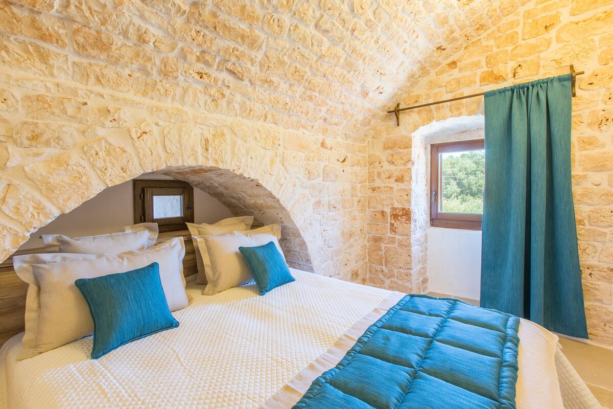 A bedroom with a stone wall, a bed with a quilt, pillows, and a window with curtains.