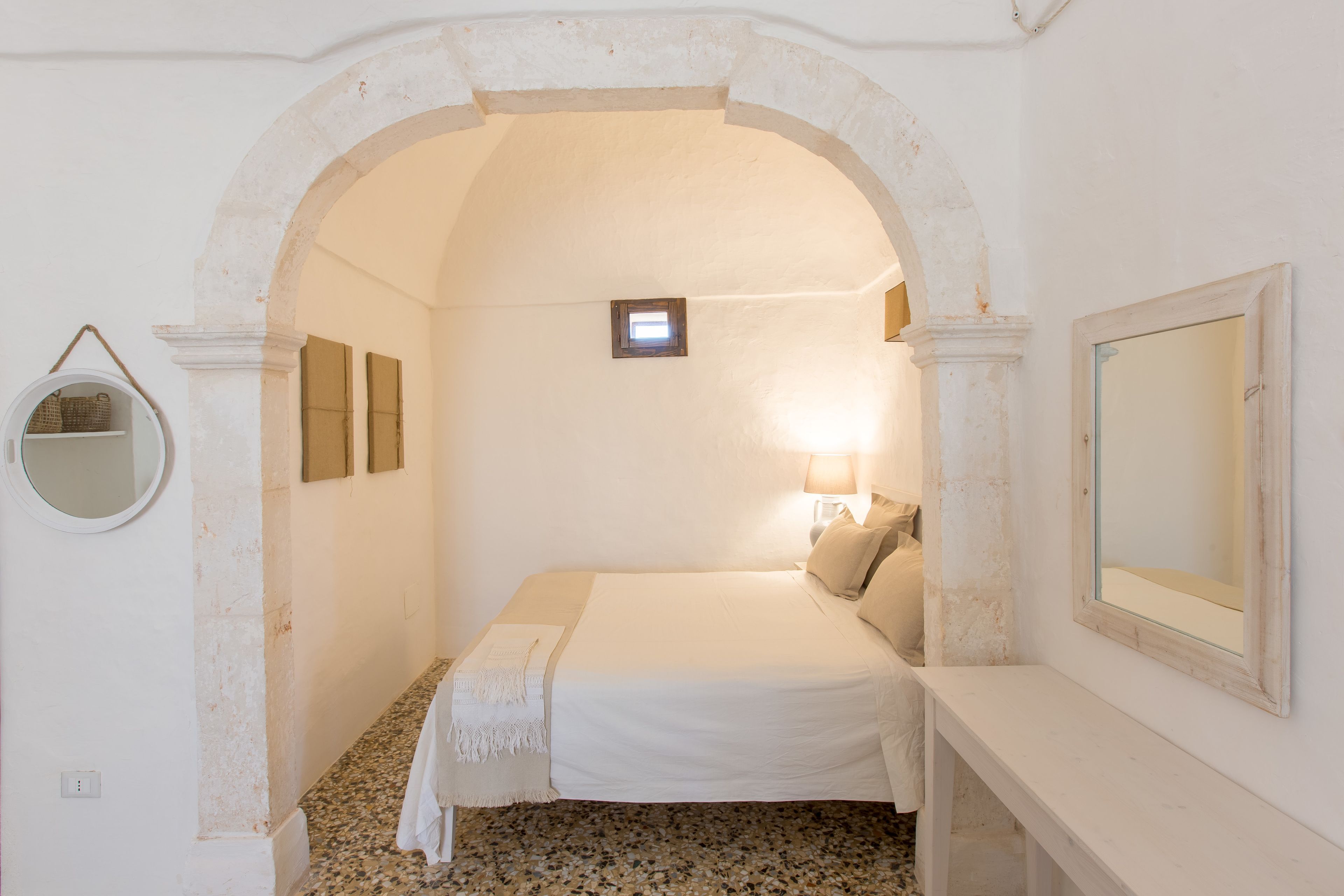 A bedroom with a bed, a bedside lamp, a mirror, and a small window.
