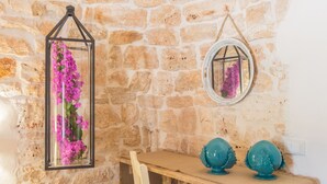 Un muro de piedra con una estructura de vidrio que contiene flores moradas, un espejo redondo y dos adornos turquesa sobre una mesa de madera.