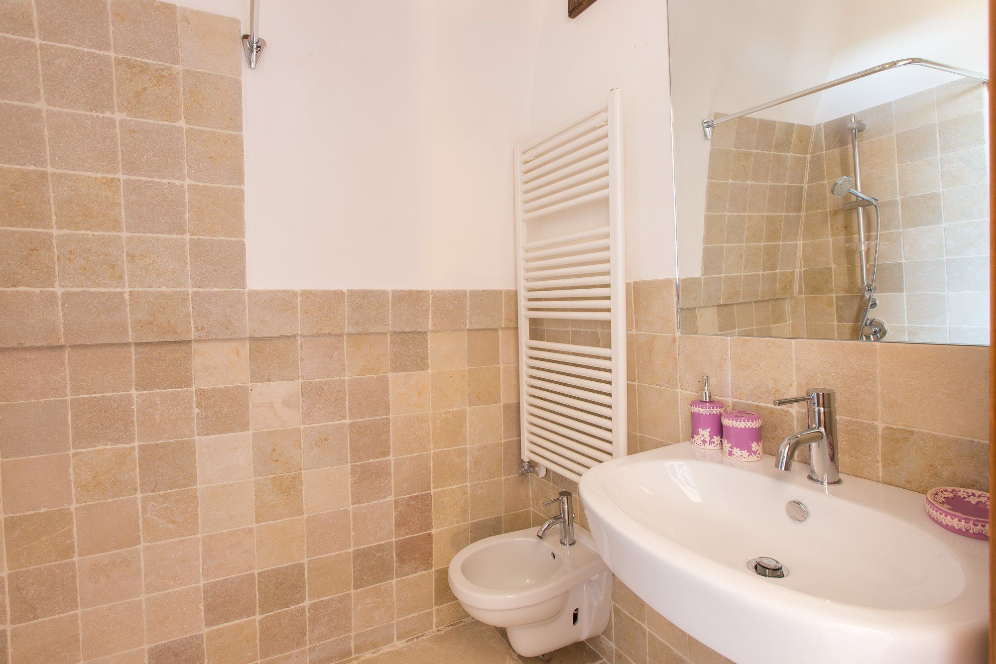 Ein Badezimmer mit beigelfarben gefliesten Wänden, einem weißen Waschbecken, einem Bidet und einem Handtuchhalter.