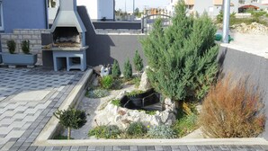 Un espace cuisine extérieur moderne, doté d’un banc en pierre et d’un barbecue encastré, entouré de parterres aménagés et de divers arbustes.