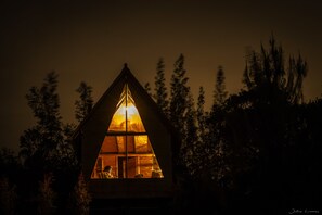 Una cabaña con techo triangular y un ventanal grande en la parte frontal, rodeada de árboles.