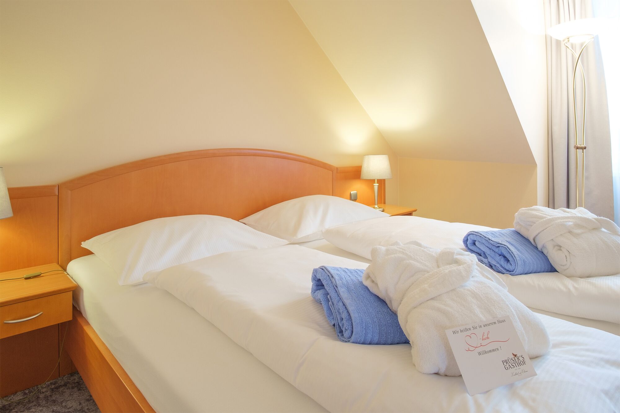 A hotel room with two beds, a wooden headboard, a nightstand with a lamp, and a window with curtains.