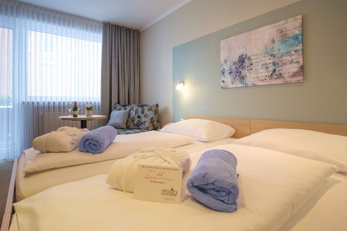 A hotel room with two beds, a small table with a bottle and glasses, a painting on the wall, and a welcome card on the bed.