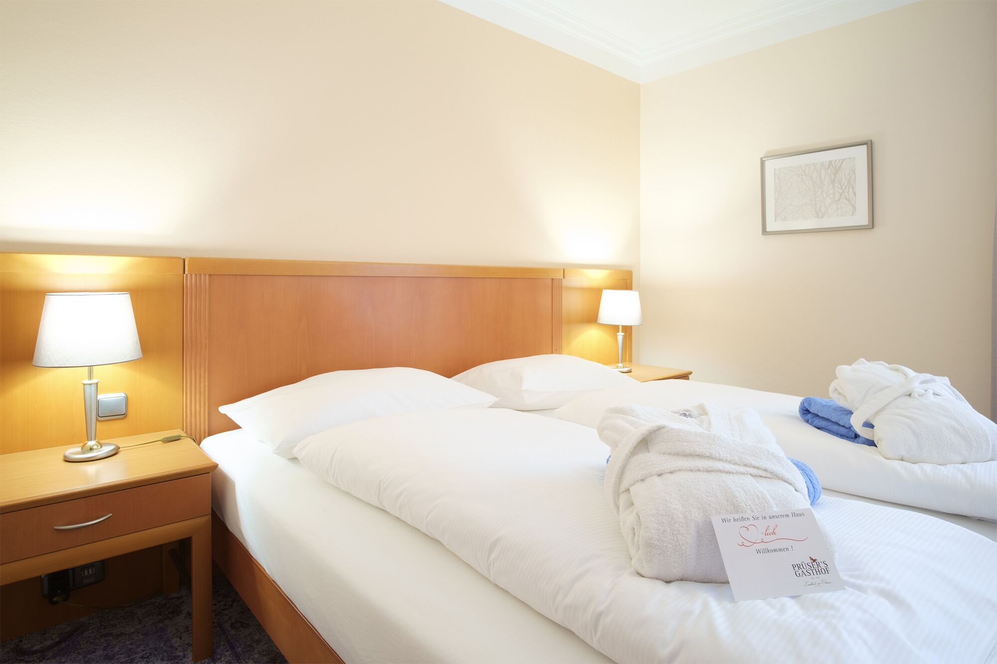 Habitación de hotel con dos camas, cabecero de madera, lámparas de noche, una mesita y una tarjeta de bienvenida.