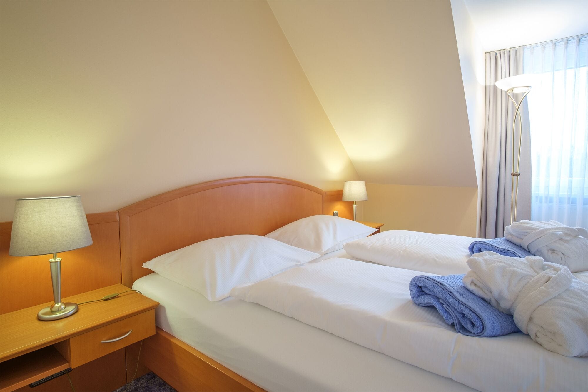 Een hotelkamer met twee bedden, een nachtkastje met een lamp en een raam met gordijnen.