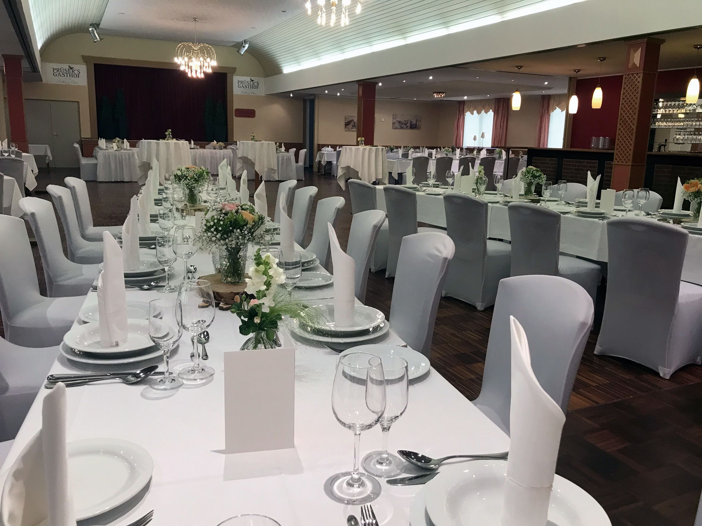 Uma sala de banquetes com várias mesas redondas preparadas para refeições, com toalhas brancas, pratos, copos e talheres bem organizados.