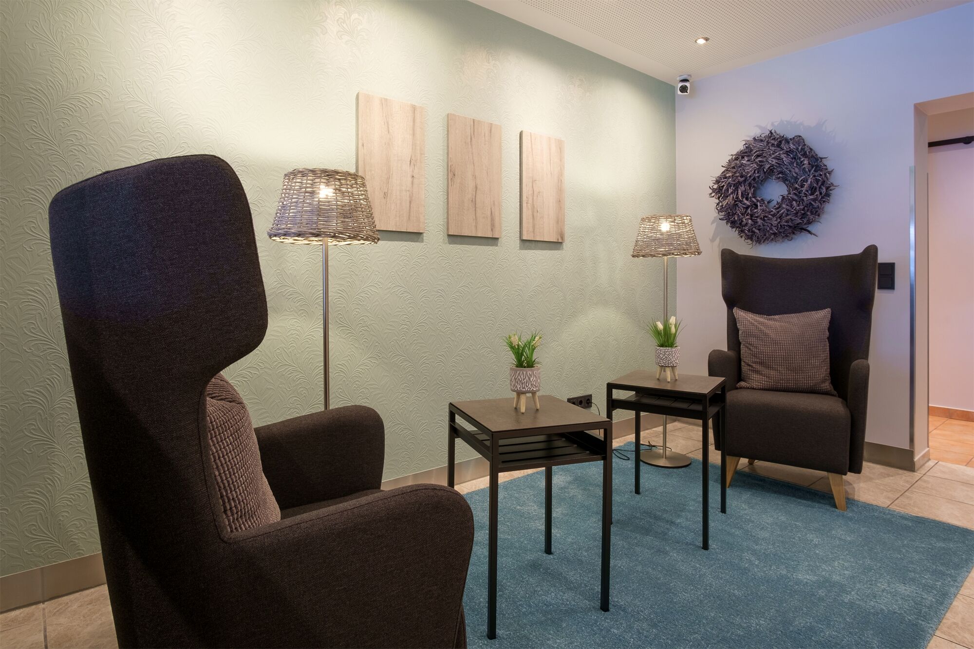 A modern waiting area with a dark brown armchair, a small table with a potted plant, and a floor lamp. There are three wooden panels on the wall and a decorative wreath.