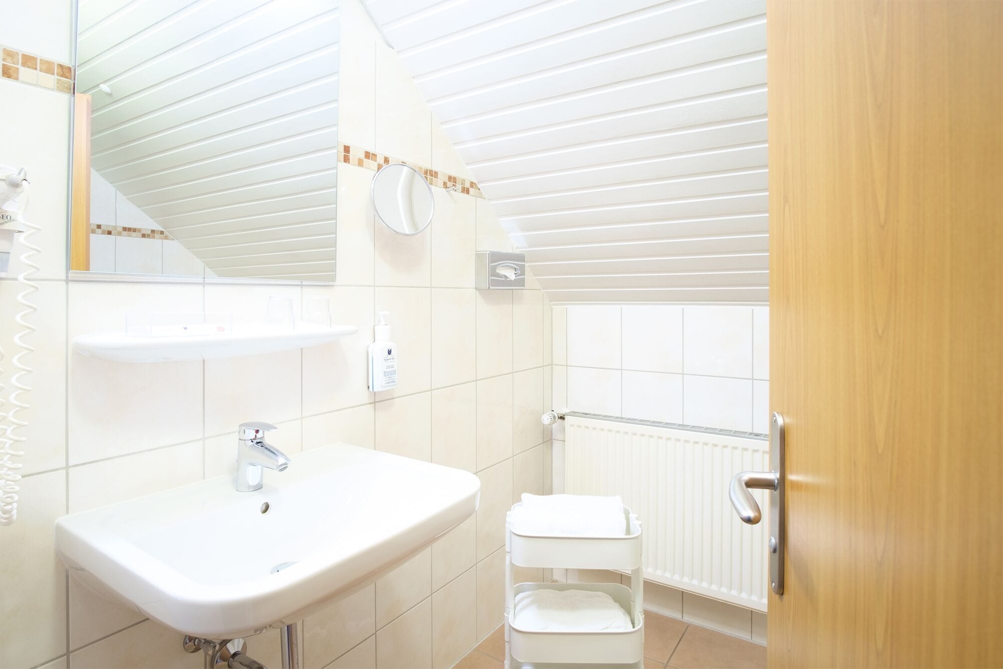 Une salle de bain moderne avec un lavabo blanc, un miroir et des toilettes. On aperçoit un porte-serviettes au-dessus du lavabo et un radiateur à l’arrière-plan.