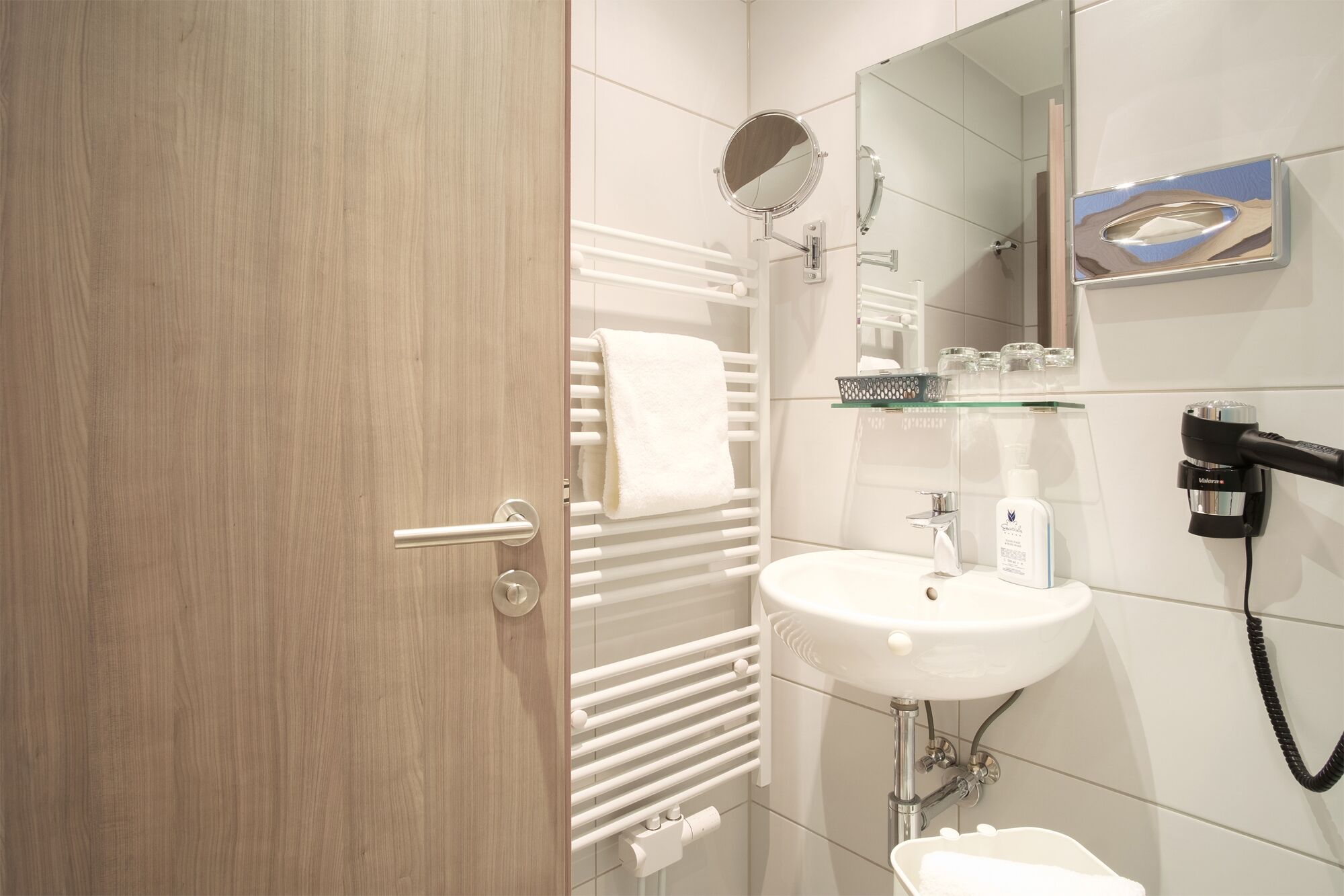 A modern bathroom with a wooden door, white tiled walls, a white sink, a mirror, a towel rack, a glass shelf with toiletries, and a hairdryer.