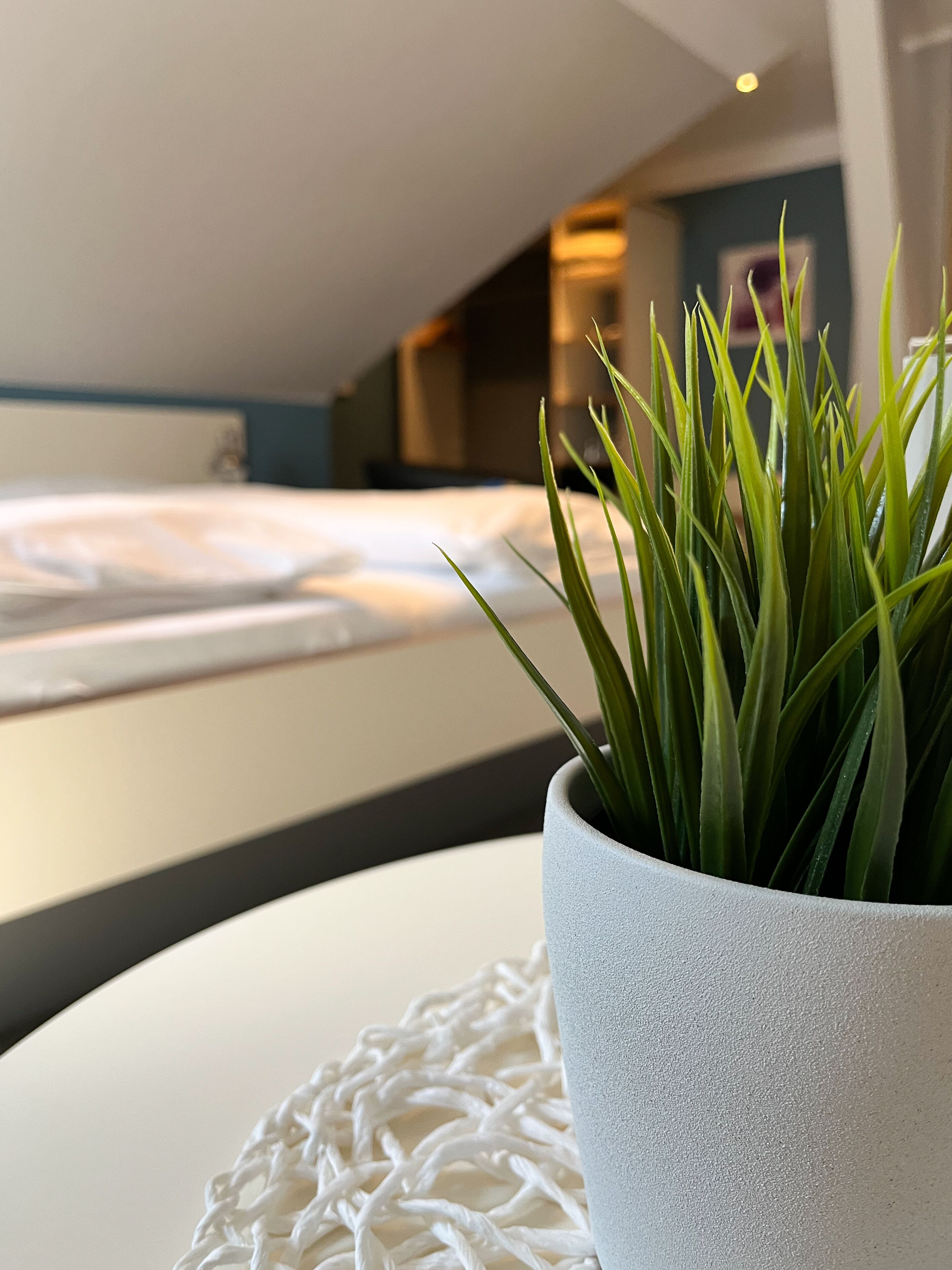Une chambre d’hôtel moderne avec un lit soigneusement fait, un tapis blanc à motifs et une plante en pot posée sur une table blanche.
