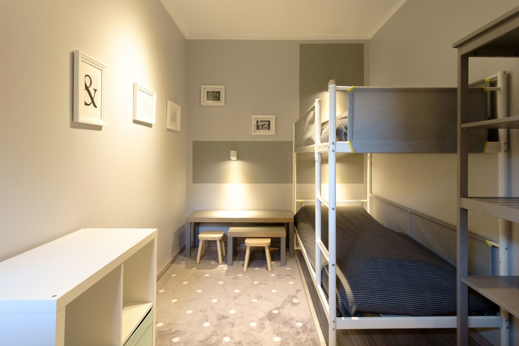 Ein Schlafraum mit Etagenbett, Schreibtisch und Stuhl, einem kleinen Hocker und einem Bett mit gestreiftem Kopfkissen.