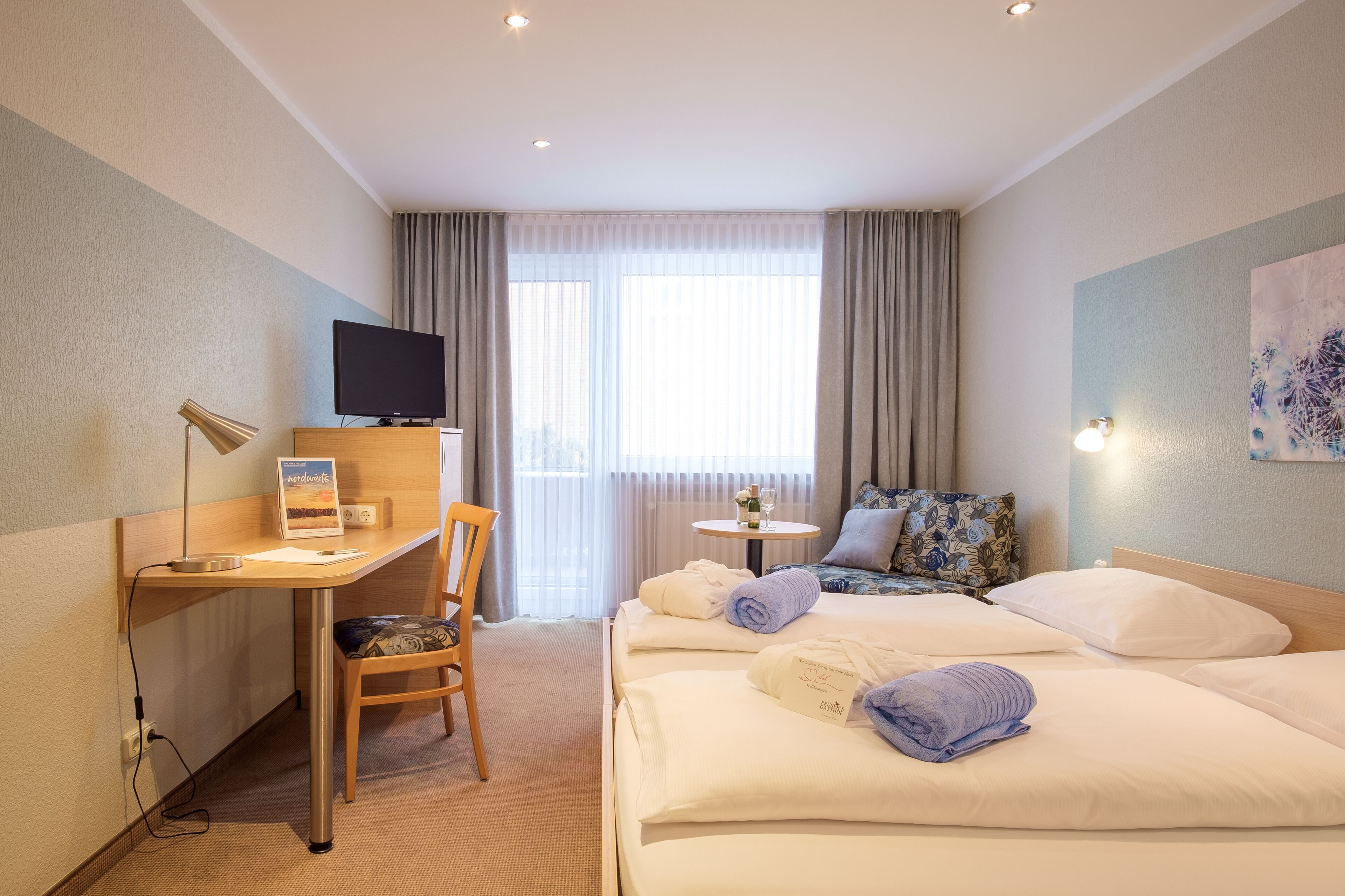 A hotel room with a desk, chair, TV, two beds, a small table, and a window with curtains.
