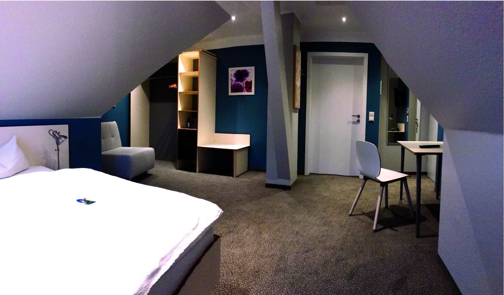 A modern hotel room with a large bed, a desk, a chair, and a bookshelf.