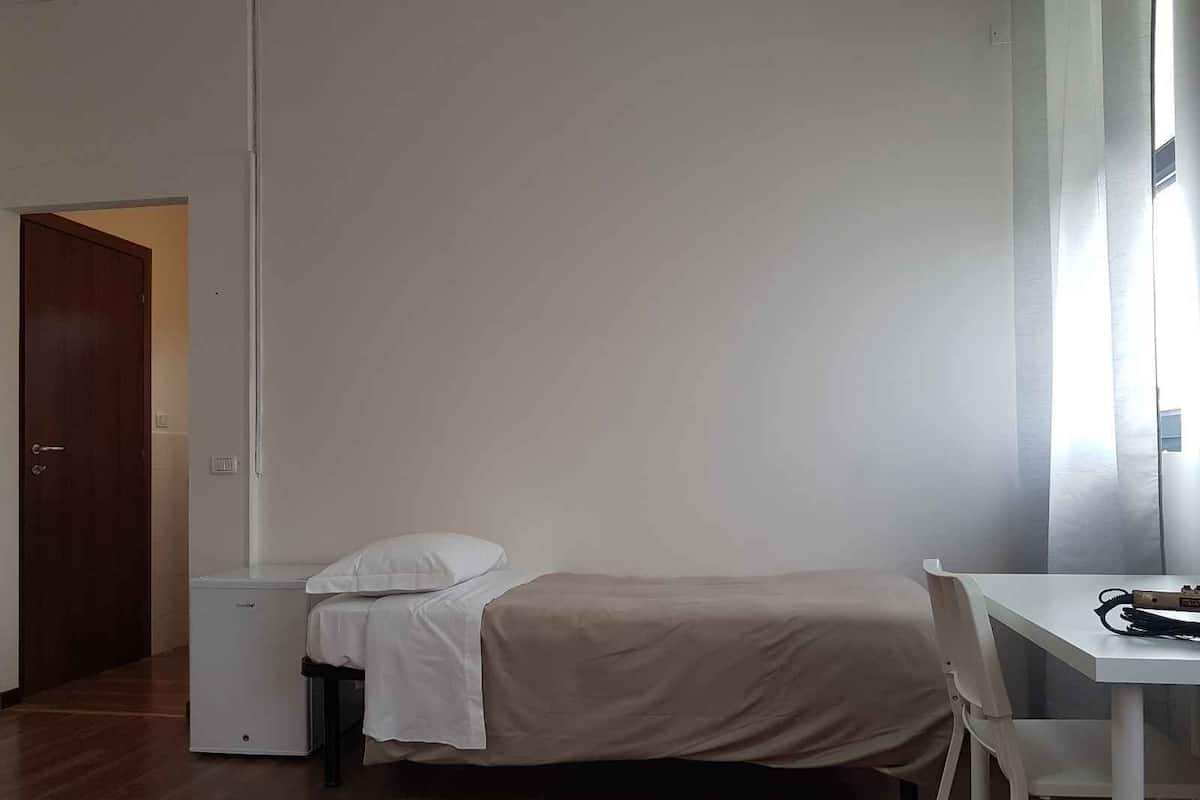 Ein kleines Zimmer mit einem Einzelbett, Holzboden, einem weißen Schreibtisch und einem kleinen Heizgerät.