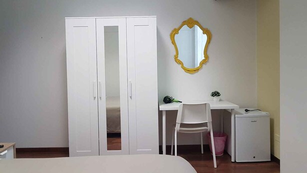 Une chambre à coucher avec un lit, une armoire blanche, un bureau avec une chaise, un petit radiateur blanc et un téléviseur fixé au mur.