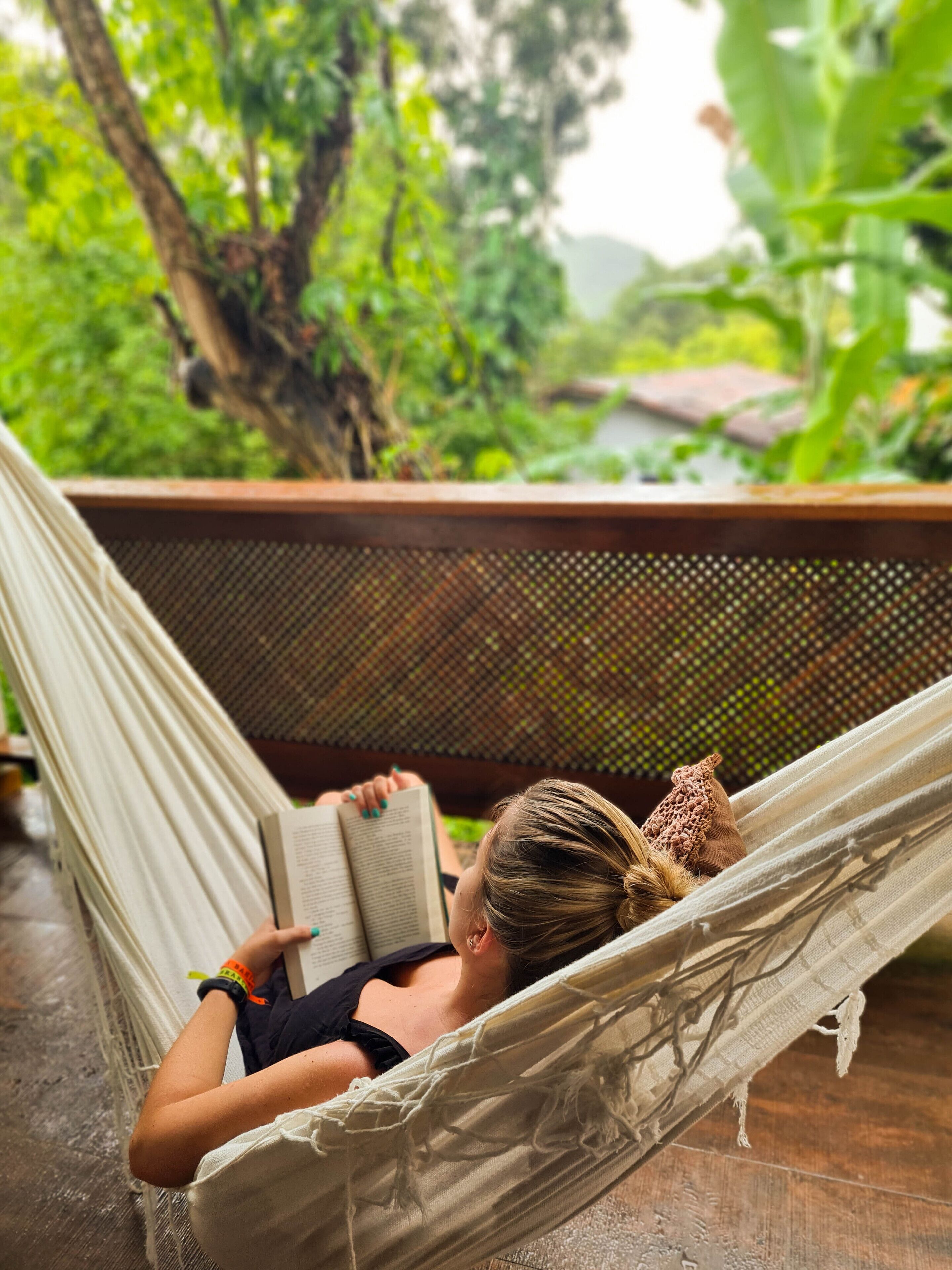 Una persona recostada en una hamaca leyendo un libro, con vistas a un follaje exuberante y una terraza de madera al fondo.