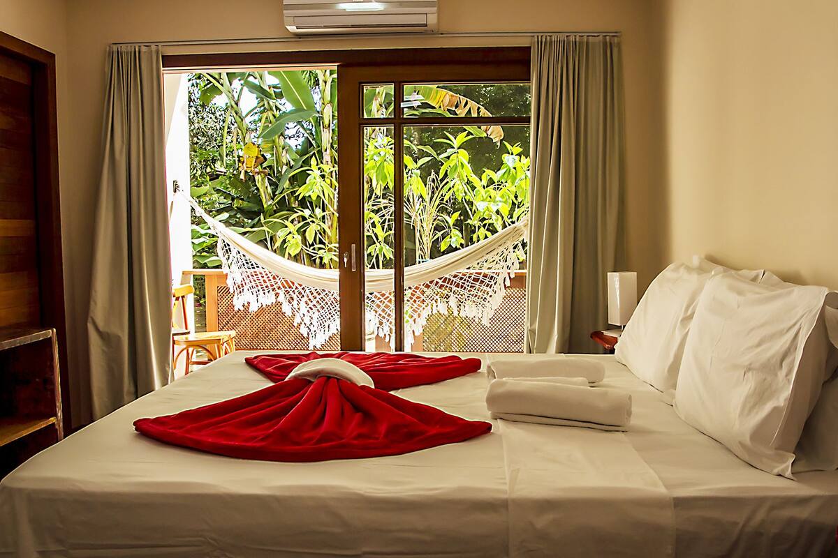 A hotel room with a bed, a hammock, and a view of greenery outside.