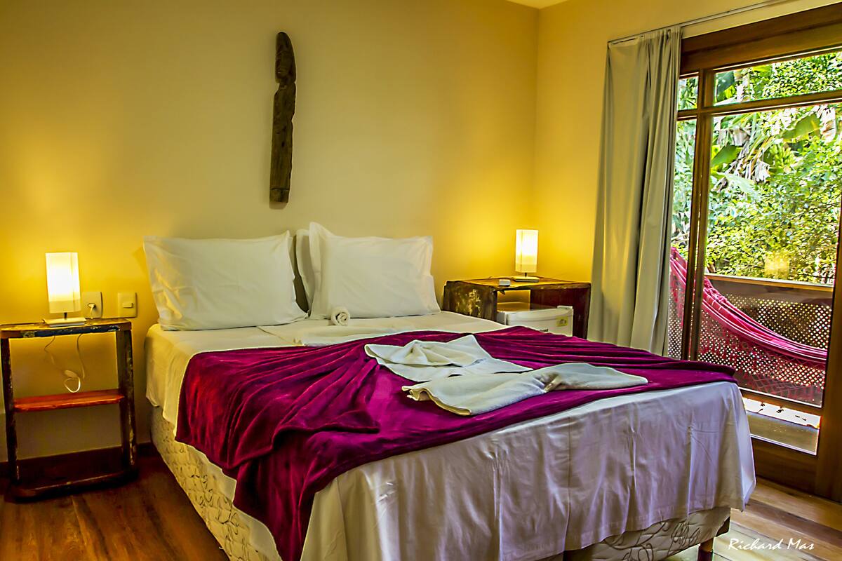 A hotel room with a bed, two bedside tables, a window with a view of greenery, and a hammock outside.
