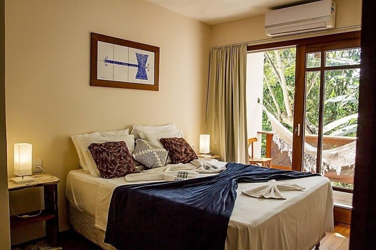 A hotel room with a bed, nightstands, a window with curtains, and a view of greenery outside.