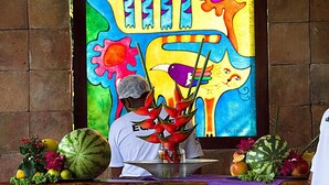 A person in a white shirt with a name tag, standing in front of a colorful abstract painting. In front of the person, there is a table with a metal tray holding a red flower arrangement, a watermelon, and various fruits and vegetables. - Pousada Timia (Cairu)