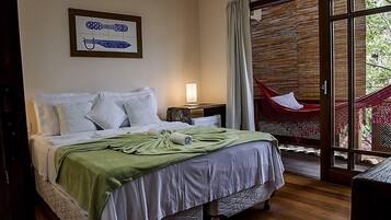 Un dormitorio con cama, almohadas, una manta verde, una mesita de noche con lámpara y una hamaca fuera de la puerta corrediza de vidrio.