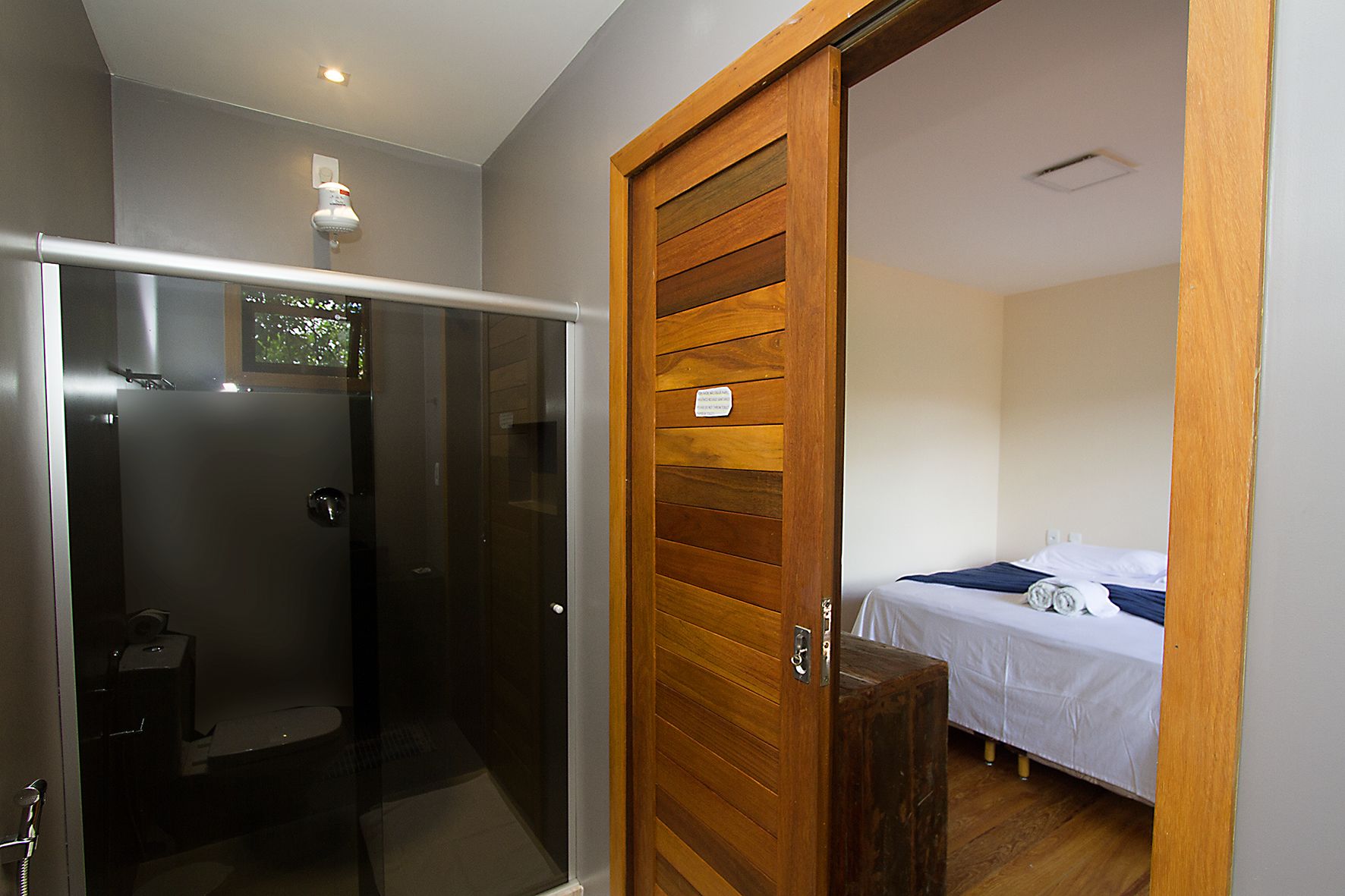 Casa de banho moderna com cabine de chuveiro em vidro e porta de madeira que dá para um quarto com cama e uma pequena mesa.