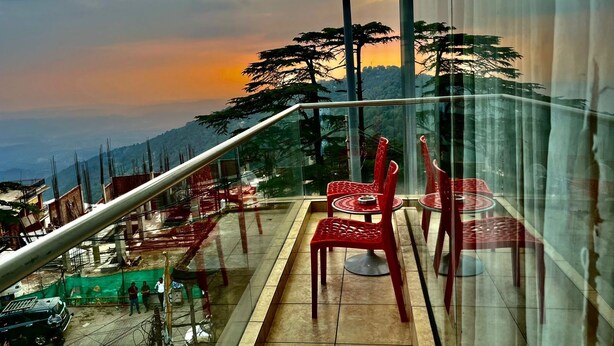 Un balcon avec des chaises rouges et une table, d’où l’on peut admirer un paysage montagneux au coucher du soleil.
