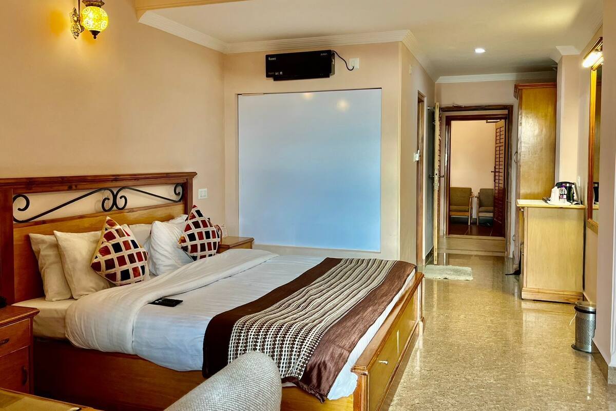 A hotel room with a bed, bedside tables, a wall-mounted TV, and a hallway leading to a sitting area.