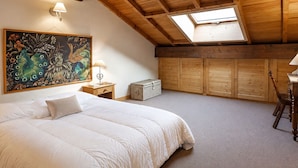 A bedroom with a large bed, a skylight, wooden beams, a painting, and a chest of drawers.