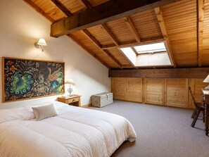 A bedroom with a large bed, a skylight, wooden beams, a painting, and a chest of drawers.