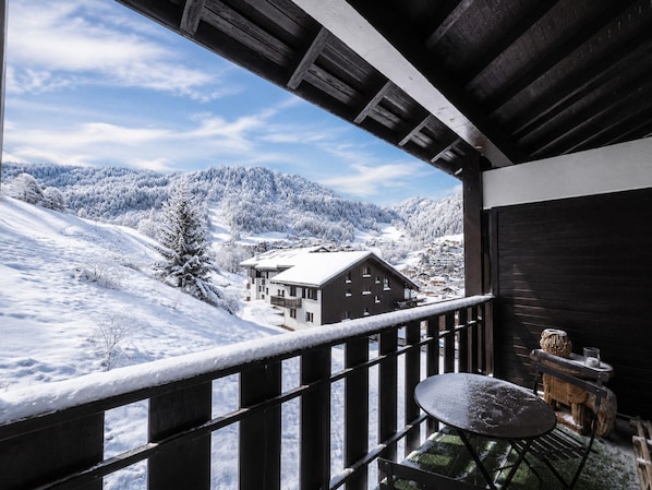 A balcony with a view of a snowy mountain landscape, a wooden building, and a table with a chair.