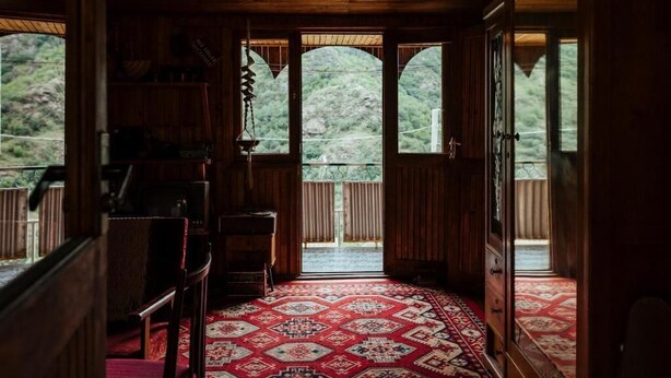 Un interior de madera con alfombra estampada, muebles de madera y ventanas amplias que ofrecen vistas al verdor exterior.