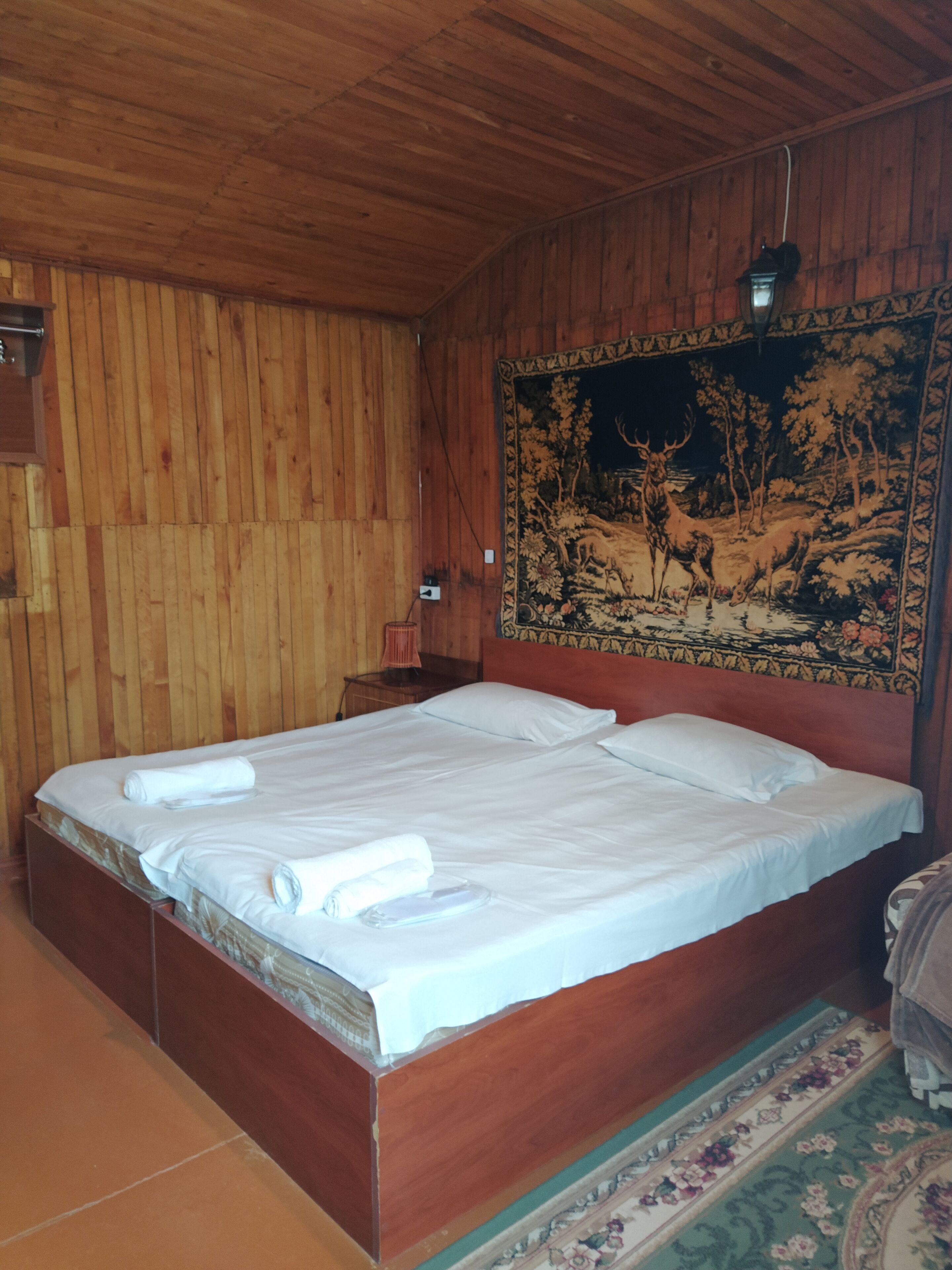 Una habitación con paneles de madera, una cama grande, un tapiz en la pared y una mesita de noche con una lámpara.