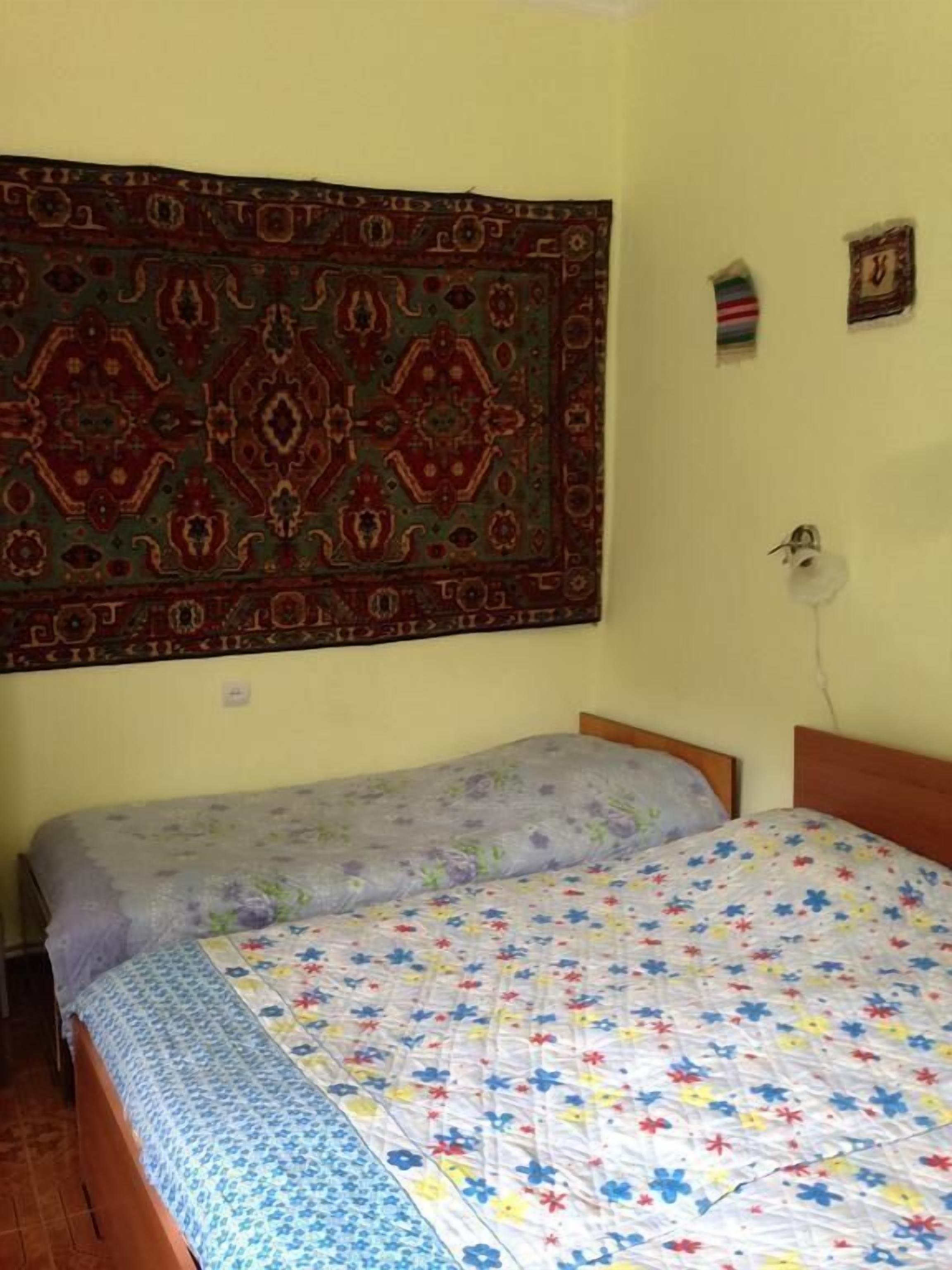 A room with two beds, one with a floral bedspread and the other with a plain one. A large patterned tapestry hangs on the wall above the beds.