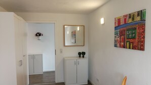Un passadís amb una porta blanca, un petit armari amb dues plantes en testos, un mirall a la paret i una obra d'art colorida.