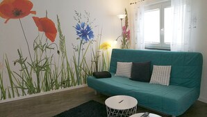 Un sofà verd marró amb coixins estampats, una taula rodona petita de color blanc i un mural de paret amb herbes altes i flors.