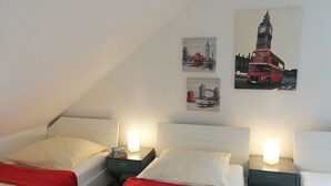 Habitació d'hotel amb dos llits, cadascun amb una manta vermella, almoinos blancs i taules de nit amb làmpades. Hi ha tres quadres penjats a la paret que representen monuments de Londres.