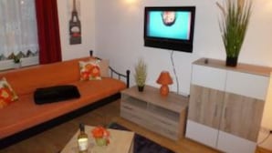 Un salon comprenant un canapé, un téléviseur fixé au mur, un meuble et une petite table sur laquelle se trouvent une bouteille et un vase avec une fleur.