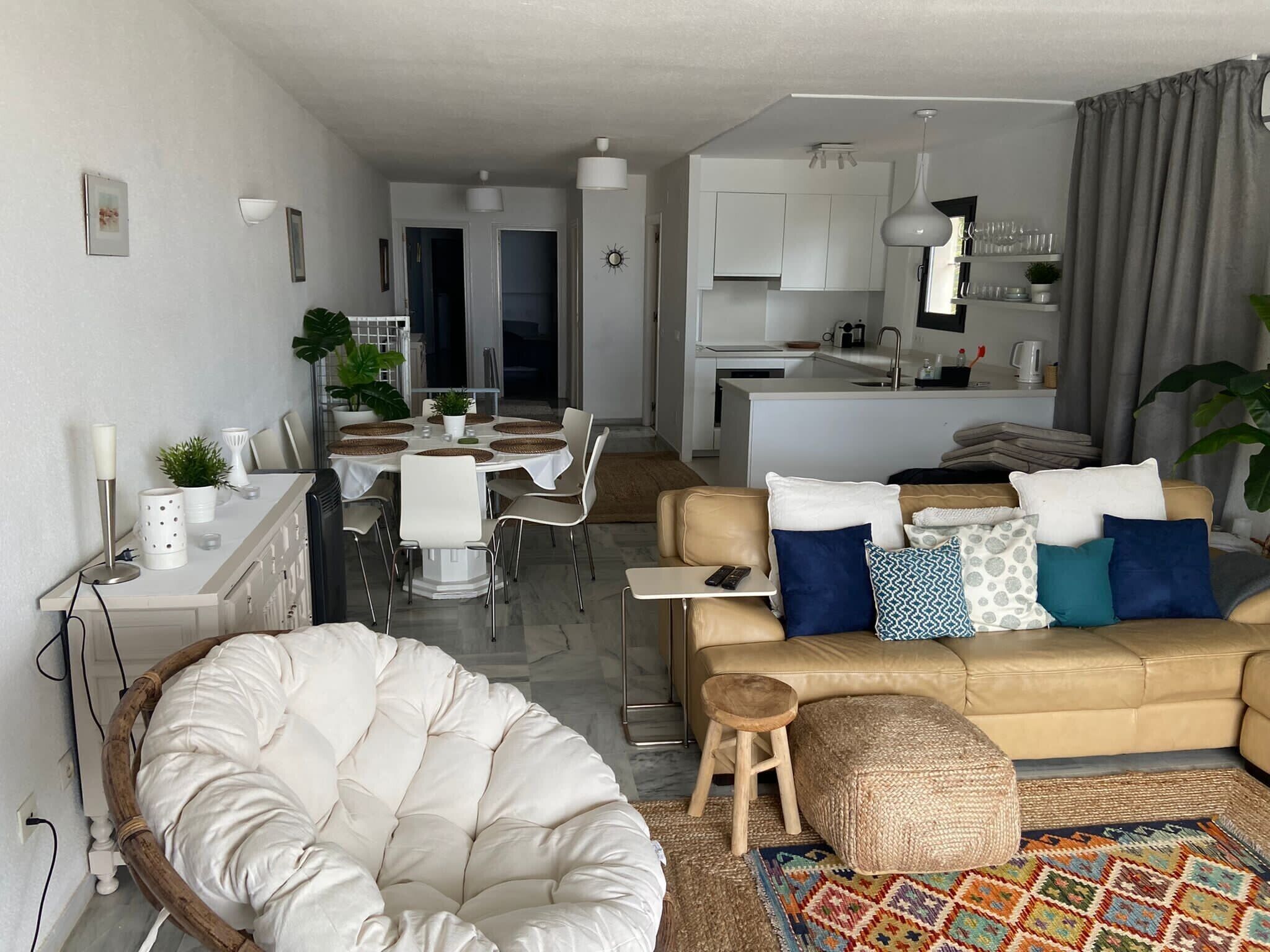 Ein modernes Wohnzimmer mit einem beigen Sofa, einem weißen Sessel und einem Essbereich mit weißem Tisch und Stühlen.