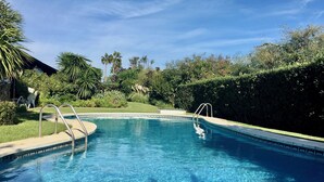 Una piscina cristalina con una escalera metálica, rodeada de exuberante vegetación y un edificio de piedra al fondo.