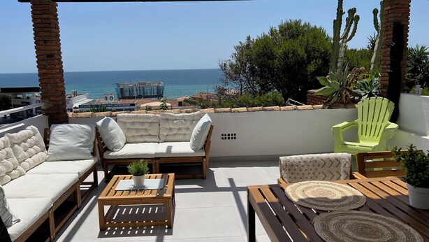 Una terraza con un sofá blanco, una mesa de madera y una silla verde, con vistas a una playa y un edificio de techo azul.