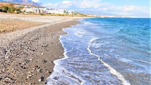 Una playa con piedras y olas, edificios al fondo y un cielo despejado.