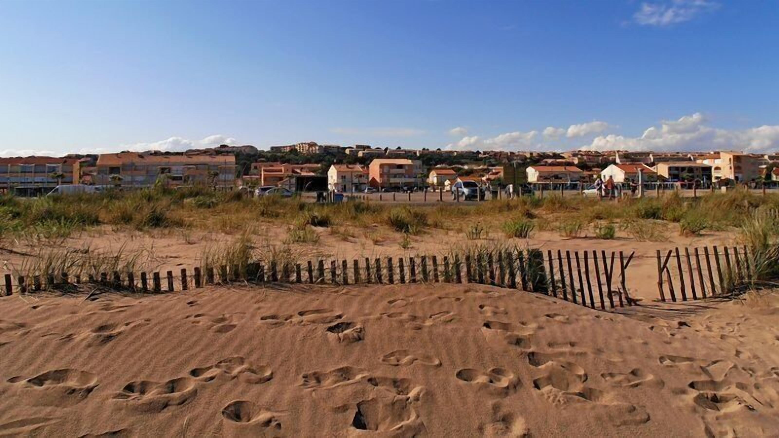 Una playa arenosa con huellas, una cerca de madera y edificios al fondo.