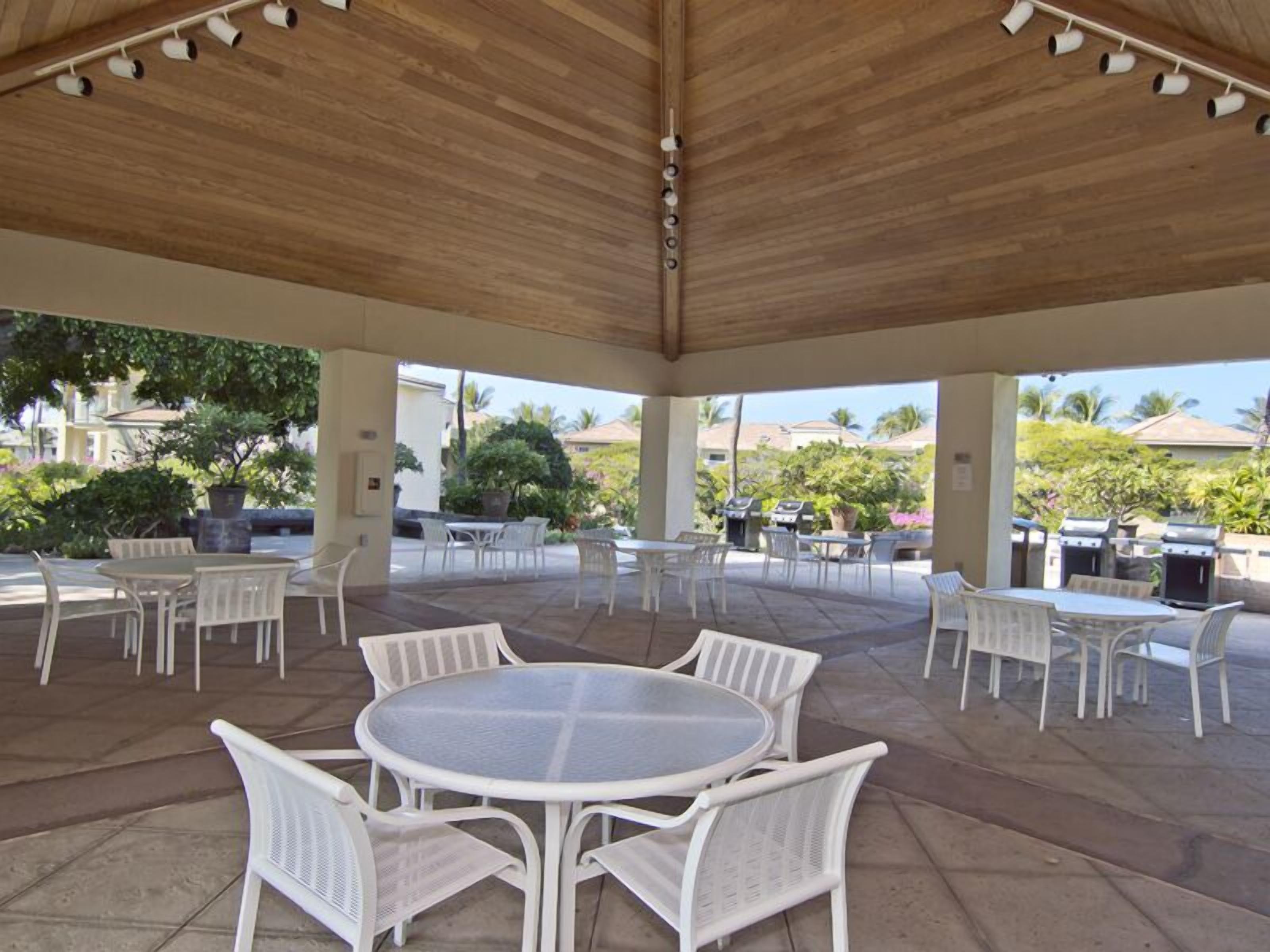 An outdoor patio area with white metal tables and chairs, a wooden ceiling, and a view of the surrounding landscape.