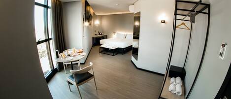 A modern hotel room with a bed, bedside table, and a small table with a chair. There is a window with curtains, a mirror, and a coat rack with a hanger.