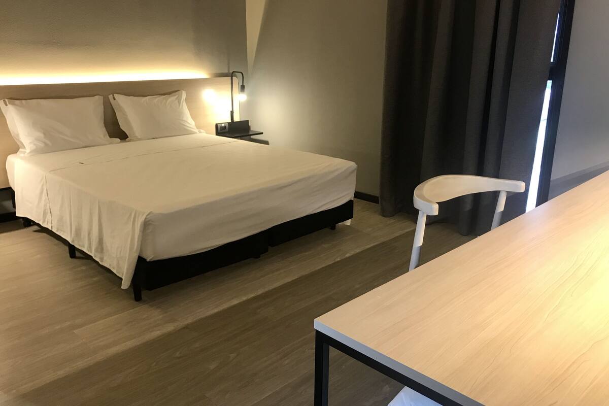 Een net opgemaakt bed met witte lakens, een nachtkastje met een lamp en een stoel in een moderne hotelkamer.