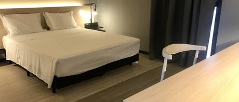 A neatly made bed with white linens, a bedside table with a lamp, and a chair in a modern hotel room.