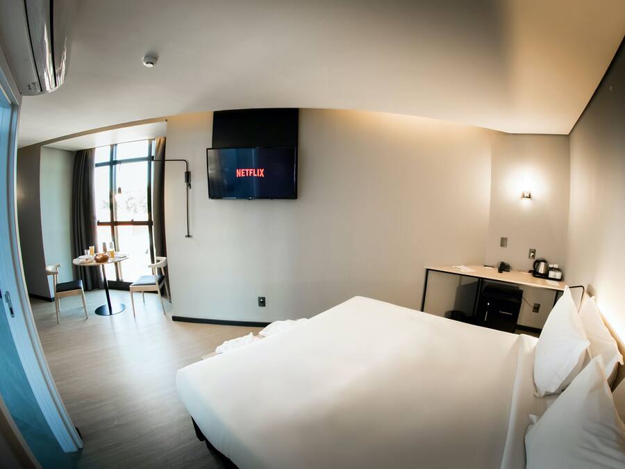 A modern hotel room with a large bed, a flat-screen TV mounted on the wall, a desk with a chair, and a small table with a bowl of fruit.