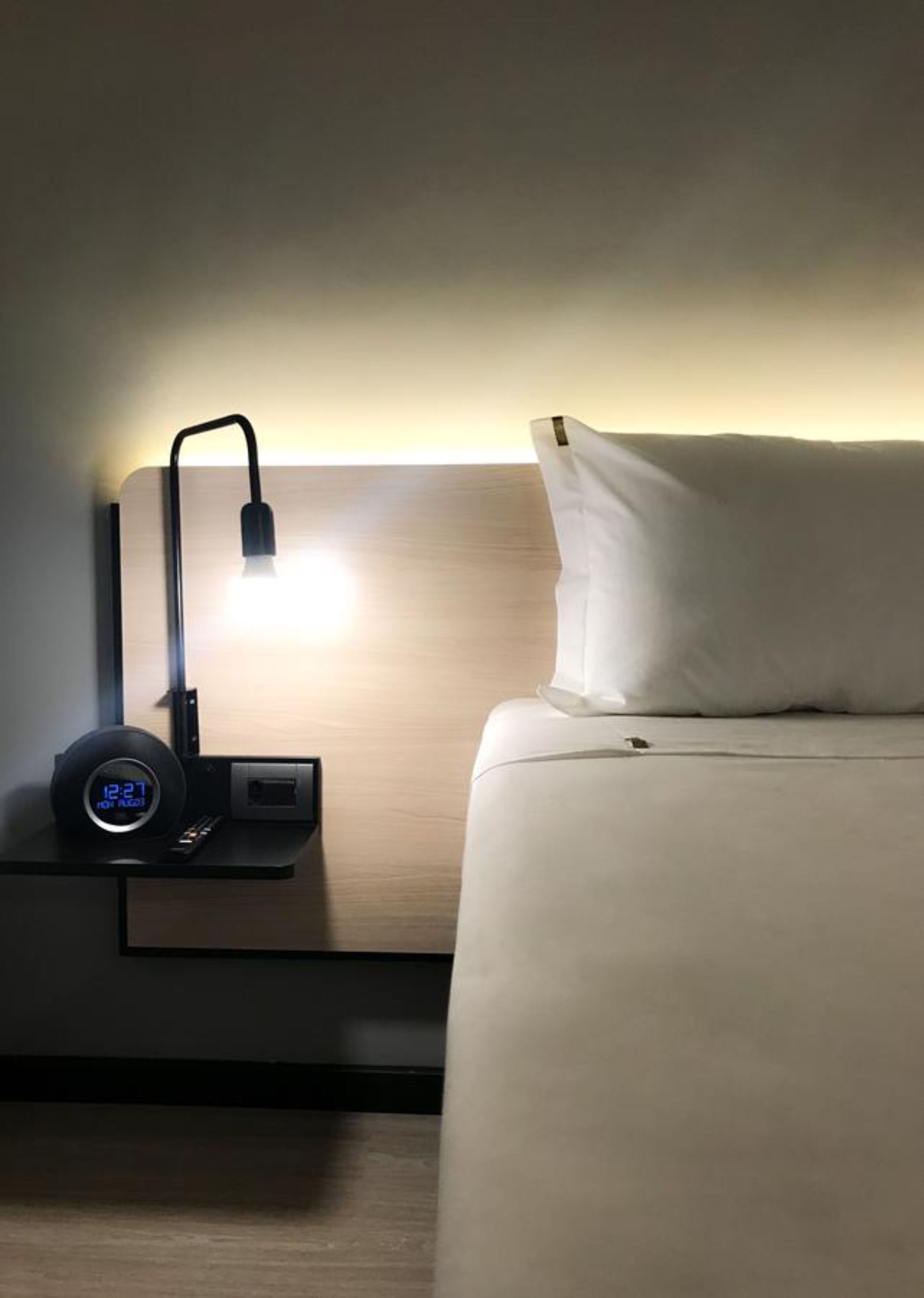 Una cama bien hecha con una almohada y una mesita de noche con un reloj digital y una lámpara.